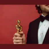 Homem vestindo smoking, visto pela metade, segurando uma estátua dourada em forma de figura humana sobre fundo vermelho, simbolizando conquista e premiação