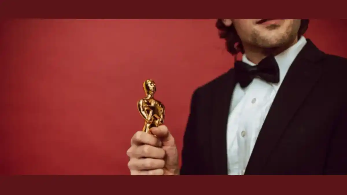 Homem vestindo smoking, visto pela metade, segurando uma estátua dourada em forma de figura humana sobre fundo vermelho, simbolizando conquista e premiação